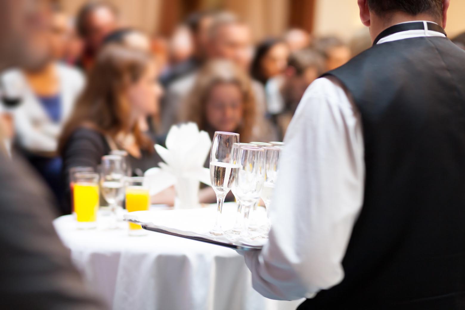 Server carrying a tray of drinks through a crowded reception.