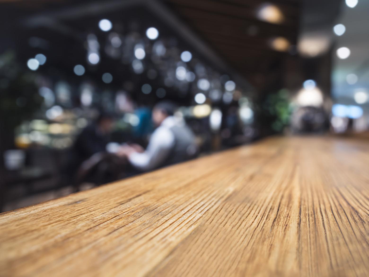 Wooden café counter with softly blurred patrons and lights beyond it.