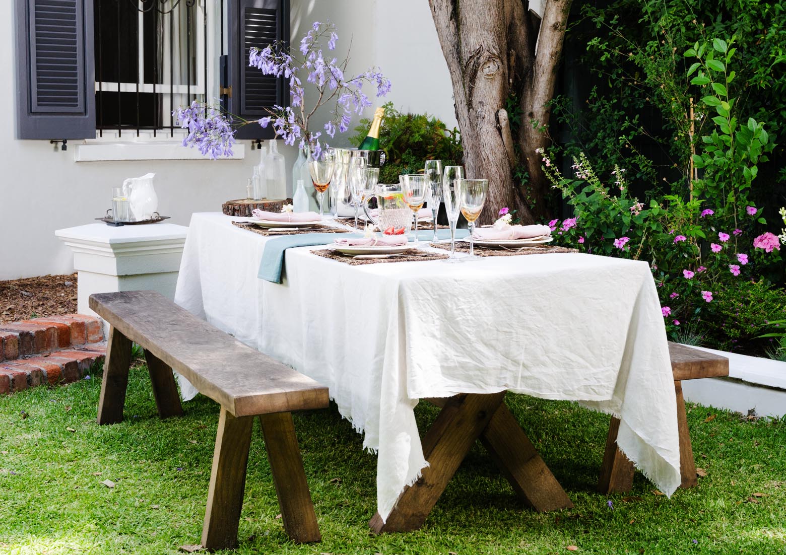 Outdoor dining table set with white linens and glassware in a garden.